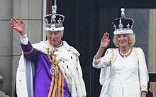 המלך צ'ארלס השלישי לבוש בבגדי ההכתרה וחבוש בכתר האימפריה הממלכתי ורעייתו המלכה קמילה חבושה בכתר המלכה מרי, מנופפים לקהל במרפסת ארמון בקינגהאם לאחר טקס הכתרתם
