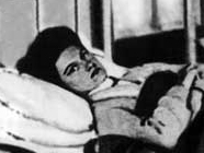 A Caucasoid woman with dark hair is lying in a hospital bed; she is looking at the camera, pictured from her mid-torso through the wall behind her head.