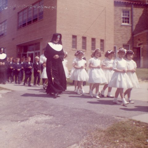 Photo two nuns walk with a line of students. The girls, at the front of the line, wear white dresses and have headbands on. The boys, in the back of the line, wear blue suits. They're on a sidewalk outside a building.