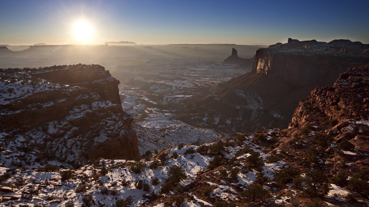 Rays of sun illuminate a sweeping overlook in Island in the Sky; highlighitng sandstone spires, canyons and vegetation dusted in snow. 