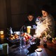 A woman adjusts an oil lamp in the dark while standing between her two sons