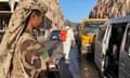 A soldier in camouflage watches as a man in protective clothing and a backpack-mounted sprayer walks along a line of vehicles at a checkpoint
