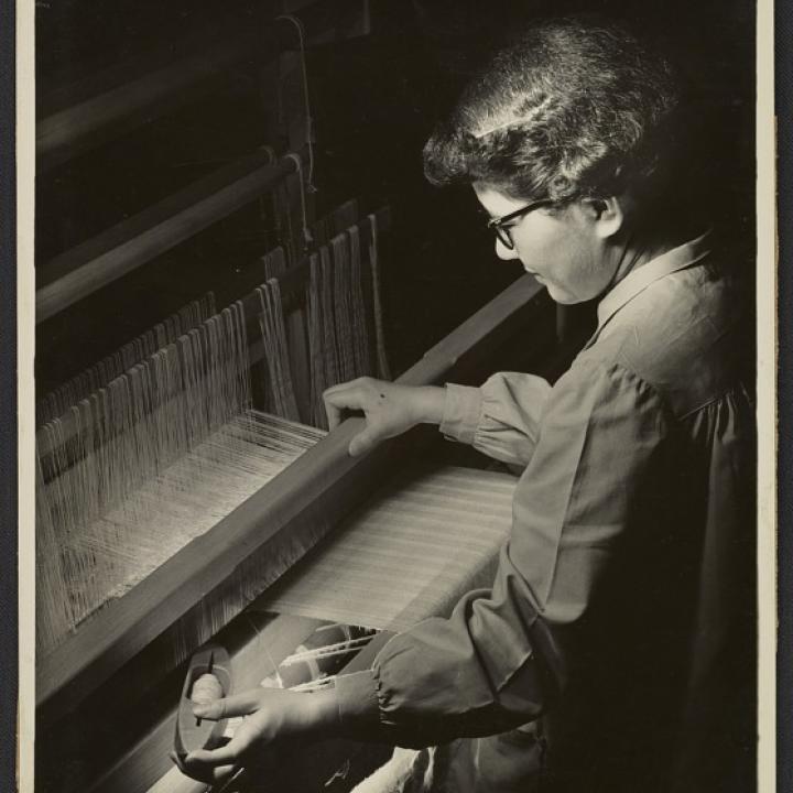 Photograph shows Kay Sekimachi weaving at a loom.