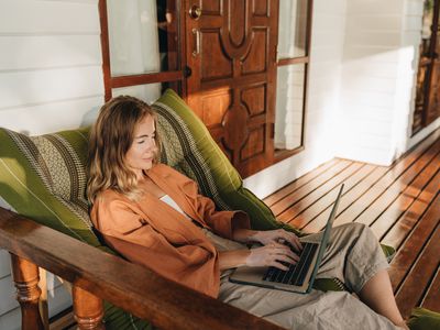 A woman relaxes in a lounge chair on a front porch. She types on the keyboard of the open laptop that rests on her lap. 