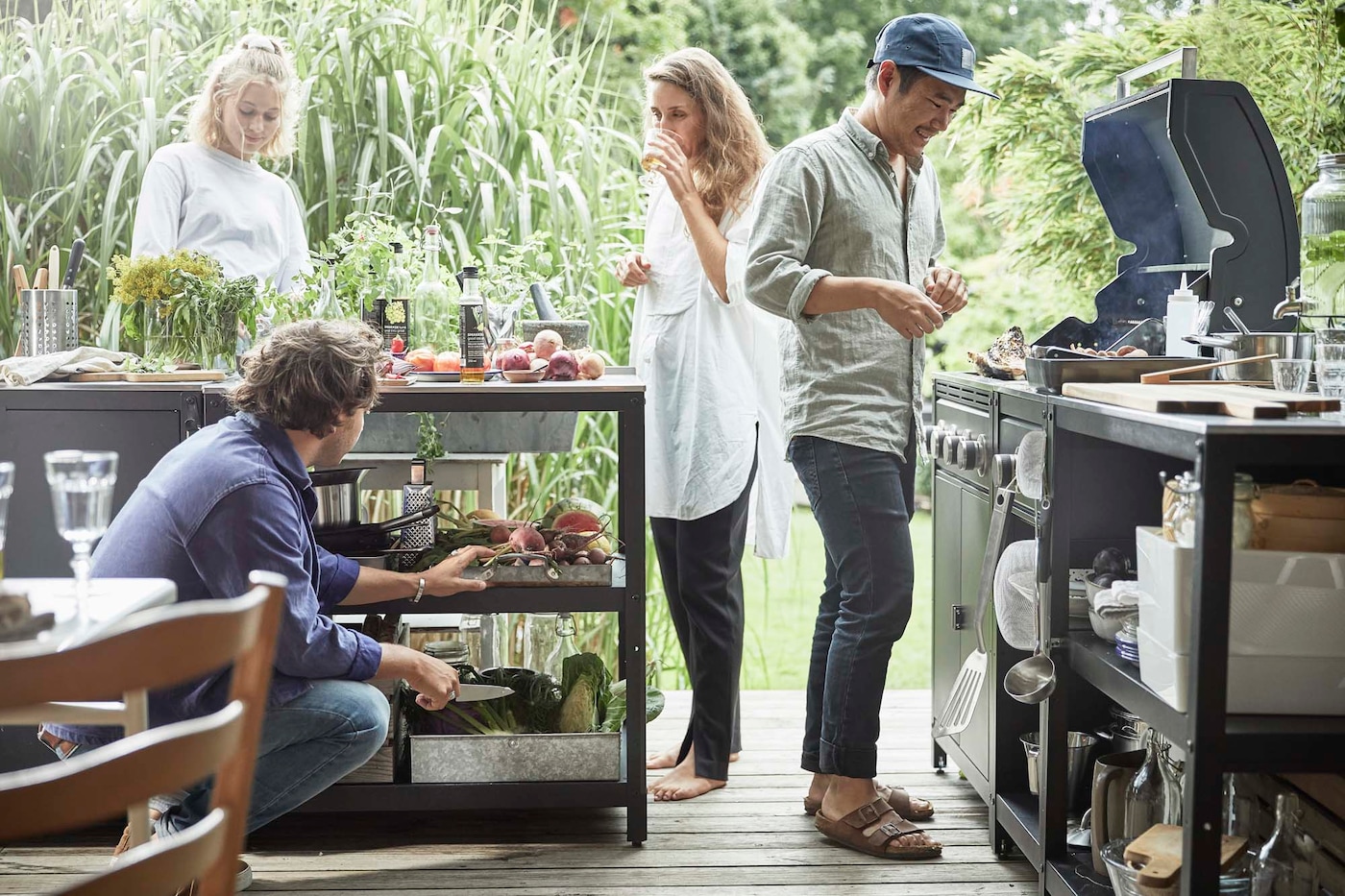 Four people grilling in an outdoor setting. There is a shelving unit with food ingredients and preparation items and a barbeque.