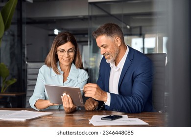 Team of diverse partners mature Latin business man and European business woman discussing project on tablet sitting at table in office. Two colleagues of professional business people working together. Foto stock
