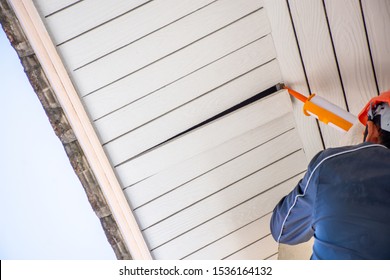 ニッカーガン、シリコーンチューブ、または手動かしめガンを手で持ち、使用して、家の外部の屋根の下の天井にある木製コルニスを修理し、固定する技術者。の写真素材