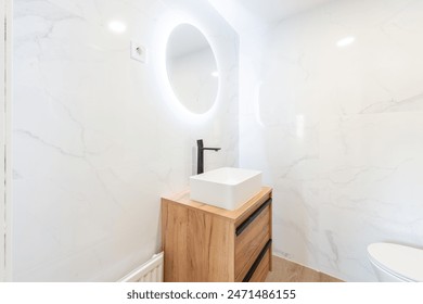 この画像では、洗練された白い大理石の壁、木製の化粧台にトレンディな長方形の洗面台、照明された円形の鏡を備えた、スタイリッシュで空いているアパートメントのバスルームを紹介しますの写真素材