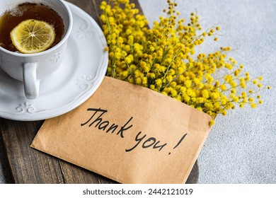 Top view of delicate cup of tea with a slice of lemon beside a cheerful bouquet of yellow Mimosa flowers and a heartfelt Thank You note, fotografie de stoc