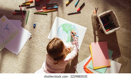 Top View: Little Girl Drawing Our Beautiful Green Planet Earth. Child Having Fun at Home on the Floor, Imagining Our Planet as a Happy Place with Clean, Sustainable Living. Cozy Sunny Day. Stock Photo