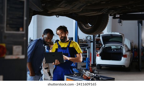 Trained mechanic and customer standing underneath car in garage using laptop to order new parts after finding defects. Employee assisting african american client by looking for components online 库存照片