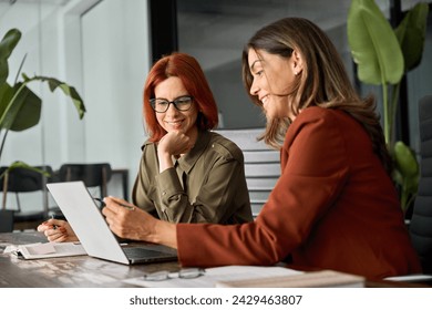 Two happy busy female employees working together using computer planning project. Middle aged professional business woman consulting teaching young employee looking at laptop sitting at desk in office