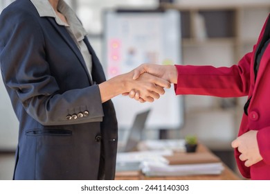 Стоковая фотография: Two women shaking hands in a business setting. The woman on the left is wearing a black jacket and the woman on the right is wearing a red jacket