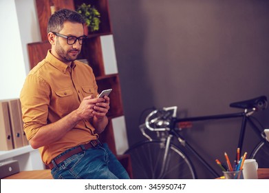 Typing business message. Confident young man looking at his smart phone while leaning at the desk in office - Φωτογραφία στοκ