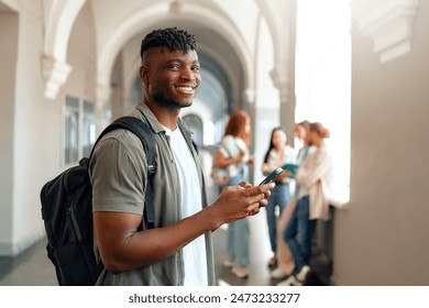 In a university hallway, a cheerful college student with a backpack is using a smartphone, surrounded by friends socializing. They epitomize the modern youthful lifestyle on campus Adlı Stok Fotoğraf