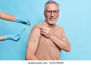 Vaccine injection against coronavirus. Pleased old man being immunized against covid 19 at hospital during visit to doctor isolated over blue wall wears plaster on arm. Vaccination for elderly people Arkistovalokuva