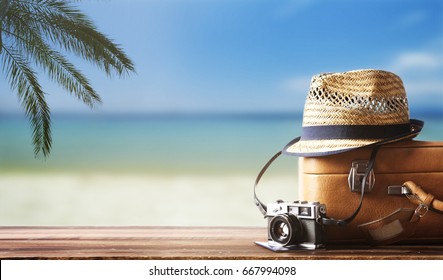Vintage suitcase, hipster hat, photo camera and passport on wooden deck. Tropical sea, beach and palm three in background. Summer holiday traveling concept design banner with copyspace. Stock Photo