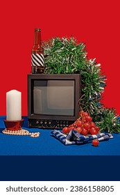 Vintage television set surrounded by an array of objects, including a bottle with a striped label, fresh grapes on a checkered cloth, a white candle and green tinsel, all set against a red backdrop Stock Photo