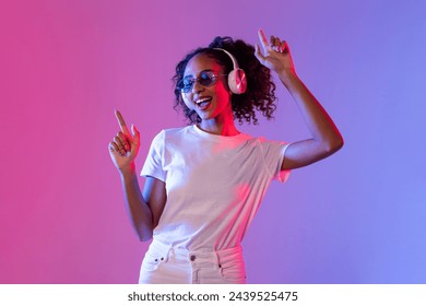 Jeune femme noire animée aux cheveux bouclés jouant de la musique dans les écouteurs sans fil et dansant sur fond rose et bleu dégradé : photo de stock