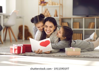 We love you. Smiling young woman getting presents from kids. Two cute twin daughters giving mom handmade greeting card. Little children lying on floor, hugging mommy and wishing her Happy Mother's Day Stock Photo