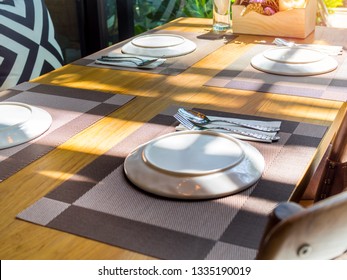 White plates, stainless steel spoons and forks on plate mats with bouquet purple flowers on wooden table. Dining table set in restaurant near glass window with sunshine. Foto Stock