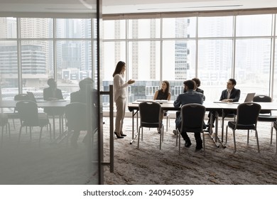 Wide shot of multiethnic business team talking at meeting table in office conference room, brainstorming in co-working space with large window. Project leader woman talking to colleagues Arkistovalokuva