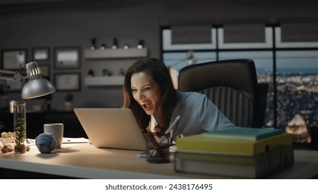 Woman winner looking laptop celebrating success in evening office. Happy boss screaming loudly enjoying business achievement. Emotional lady gesturing hands exciting by good news in dark cabinet  Stock fotografie
