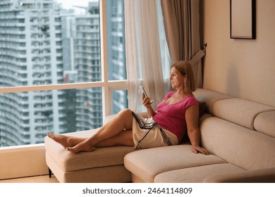 Woman looking at her phone at big window with city view. Female enjoying calm quiet time with social media. People read and relax. Modern apartment in high rise building. Foto Stock