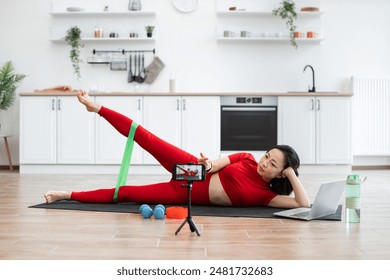 Woman in red workout clothes exercising with resistance band at home using laptop for online fitness class. Equipment includes dumbbells, yoga mat, water bottle. Bright, modern kitchen background. 库存照片