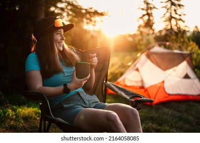 女性のテントの外の椅子に座って。森の中でキャンプする夕日。レクリエーション野外活動。キャンプでキャンプ用具と夕食を調理。夏の旅行屋外アドベンチャーの写真素材