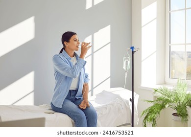 Woman with sterile tube in arm sitting by pole with IV bottle at medical center, drinking water and receiving vitamin therapy infusion to boost body immune system, preserve beauty or fight hangover Foto stock