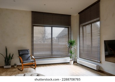 Wooden blinds on large windows in the interior. Living room with armchair and houseplants near windows with wood blinds. Motorized jalousie in the smart house. Foto stock