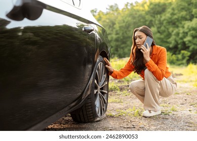Worried young European woman talking on cellphone and checking her car flat tire on the side of the highway, calling for help