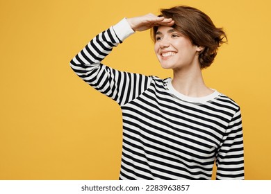 평범한 줄무늬의 검은색과 흰색의 셔츠를 입은 젊은 여성은 멀리 보이는 이마를 손으로 잡는 것이 평범한 노란색 배경스튜디오 초상화에 멀리 떨어져 있는 것처럼 보인다. 라이프스타일 개념 스톡 사진