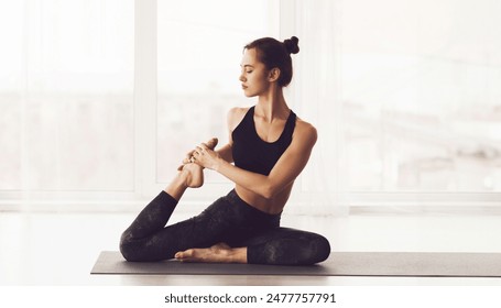 A young woman is depicted sitting on a yoga mat in a well-lit studio. She is dressed in black athletic wear and is performing a yoga pose, maintaining a focused yet serene expression Foto de stock