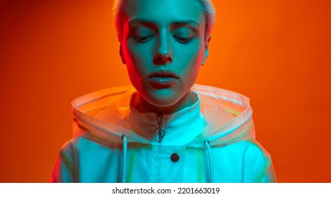Young woman in futuristic outfit with short hair standing under turquoise neon light against orange background 库存照片