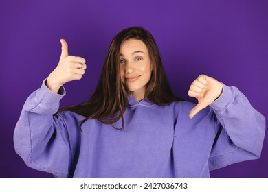 Mujer joven con capucha púrpura haciendo buen-mal signo aislado sobre fondo violeta. La chica elige, o toma una decisión con el pulgar hacia arriba o hacia abajo, guste o no, sí o no. Foto de stock