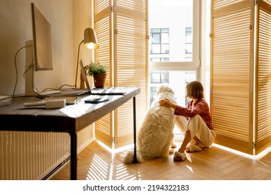 Young woman sits with her dog by the window blinds and looks away in cozy and sunny living room of modern apartment in beige tones. Quarantine, loneliness and life at home concept Foto stock