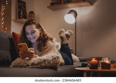 A young woman spends a cozy winter evening at home lying on the sofa looking at her smartphone. Winter holidays, Christmas and online surfing  Foto Stock