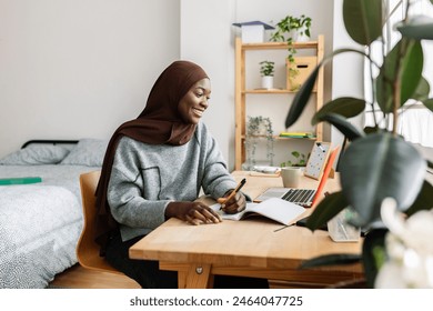 Young adult african student woman taking notes while using laptop computer at home. Millennial black female in muslim headscarf learning online listening virtual video call. Education concept. Stock-foto