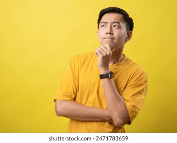 A young Asian man in a yellow shirt stands thoughtfully with his hand on his chin and arms crossed, against a solid yellow background Foto stock