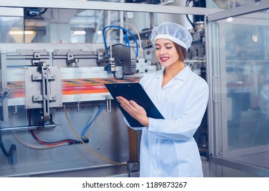A young beautiful girl in white overalls makes notes in a tablet on the background of equipment of a food processing plant. Quality control in production. स्टॉक फ़ोटो