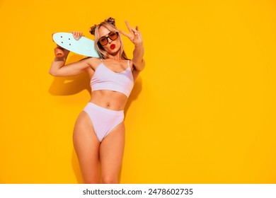 スタジオで黄色い壁の近くにポーズ若い美しい笑顔の女性。水着の水着にセクシーモデル。赤い唇を持つ陽性の女性。ペニーのスケートボードを持つ。幸せと陽気な、平和の兆候を示していますの写真素材