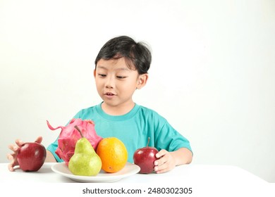 A young boy in a turquoise shirt examines a plate of colorful fruits, including dragon fruit, pear, orange, and apples. Ideal for promoting healthy eating, nutrition education, and children's health. – Ảnh có sẵn