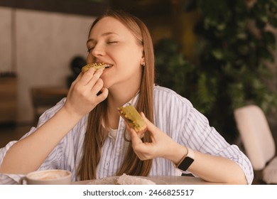 Young blonde woman eating eclair sitting in cafe. Girl bite piece of croissant look joyful at restaurant. Cheat meal day concept. Woman is preparing with appetite to eat eclair. Enjoy pistachio bakery Stock-foto