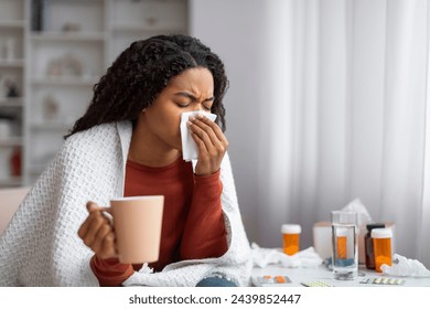 Young black woman suffering cold, blowing her nose and holding cup of tea, sick african american female sitting at home surrounded by medicines, showing symptoms of flu, copy space: stockfoto
