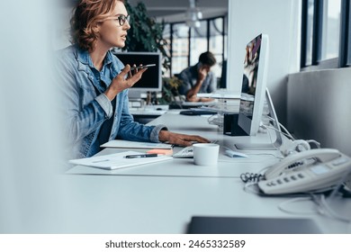 Young businesswoman talking on speaker phone and working on computer. Young woman sitting in front of monitor with smart phone. स्टॉक फ़ोटो