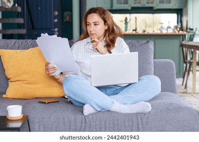 Стоковая фотография: Young freelancer woman get over paperwork while working from living room, she is sitting on the sofa with laptop in her lap