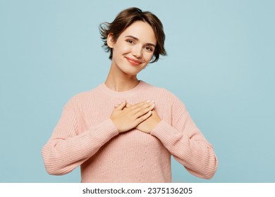 Young grateful thankful smiling happy woman wear beige knitted sweater casual clothes put folded hands on heart isolated on plain pastel light blue cyan background studio portrait. Lifestyle concept Foto Stock