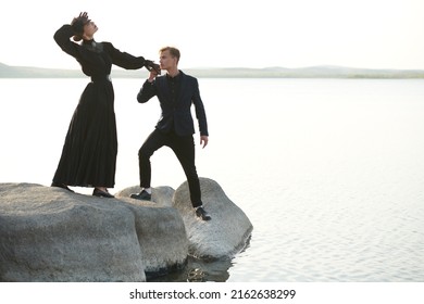 Стоковая фотография: Young handsome man kisses hand of a beautiful young woman standing on the seacoast at sunset. Historical romance novel. Loving  relationship. 19th century style.